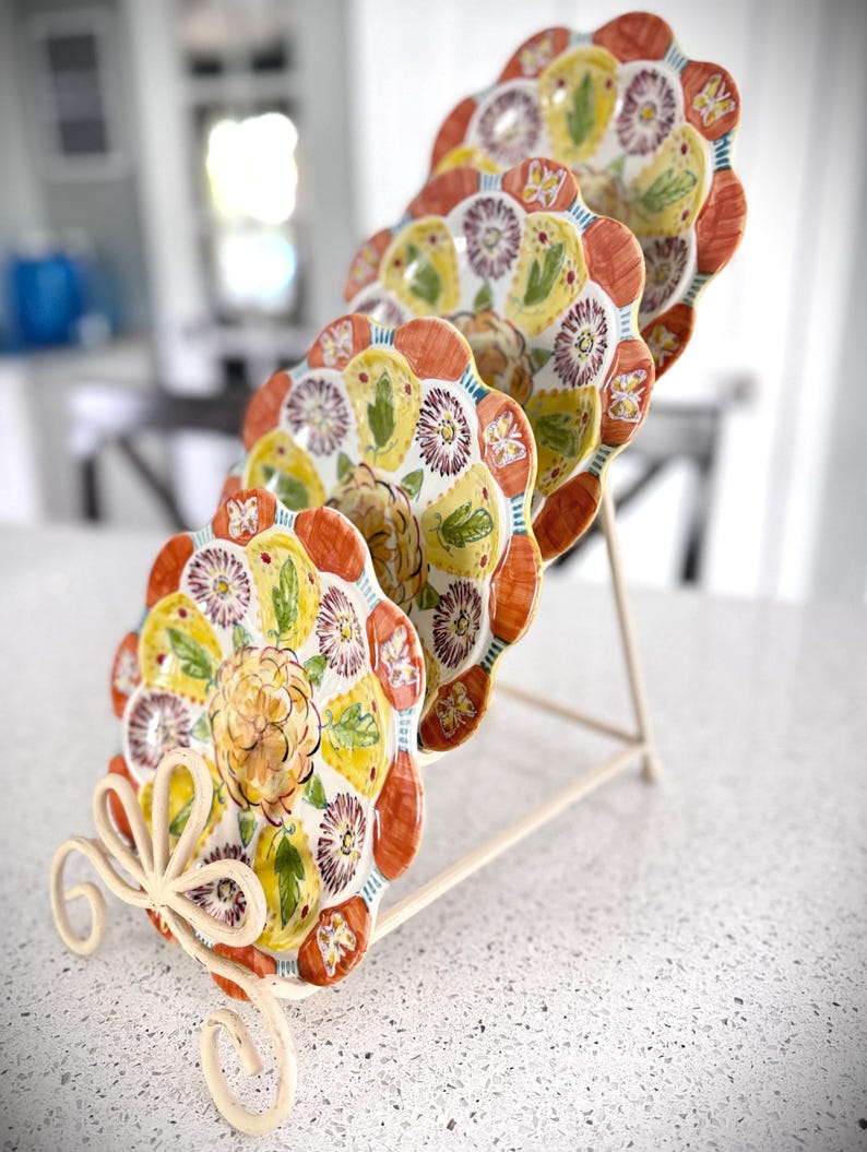 May include: A set of decorative plates with a floral design, displayed on a cream-colored metal stand. The plates feature a scalloped edge with orange, yellow, and purple floral patterns. The stand has a decorative, scrolled base.