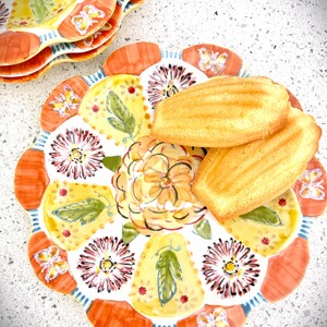 May include: A stack of decorative plates with a floral and butterfly design. The plates have a scalloped edge with orange, yellow, and white accents. Two golden-brown madeleines are on top of one plate.