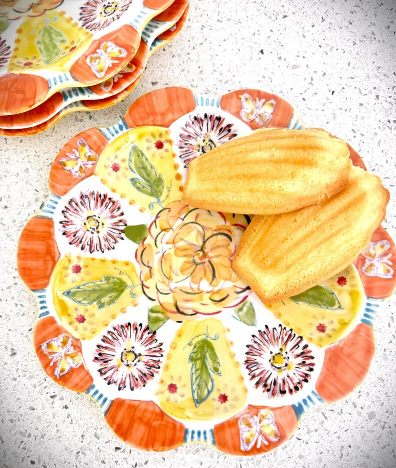 May include: A stack of decorative plates with a floral and butterfly design. The plates have a scalloped edge with orange, yellow, and white accents. Two golden-brown madeleines are on top of one plate.