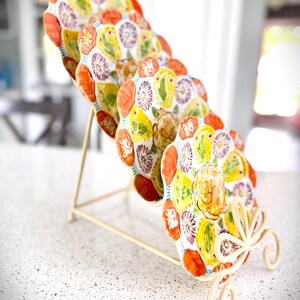 May include: A set of six decorative ceramic plates with a floral design, displayed on a cream-colored metal stand. The plates feature a white background with yellow, orange, and purple floral patterns. The stand has a decorative, scrolled base.