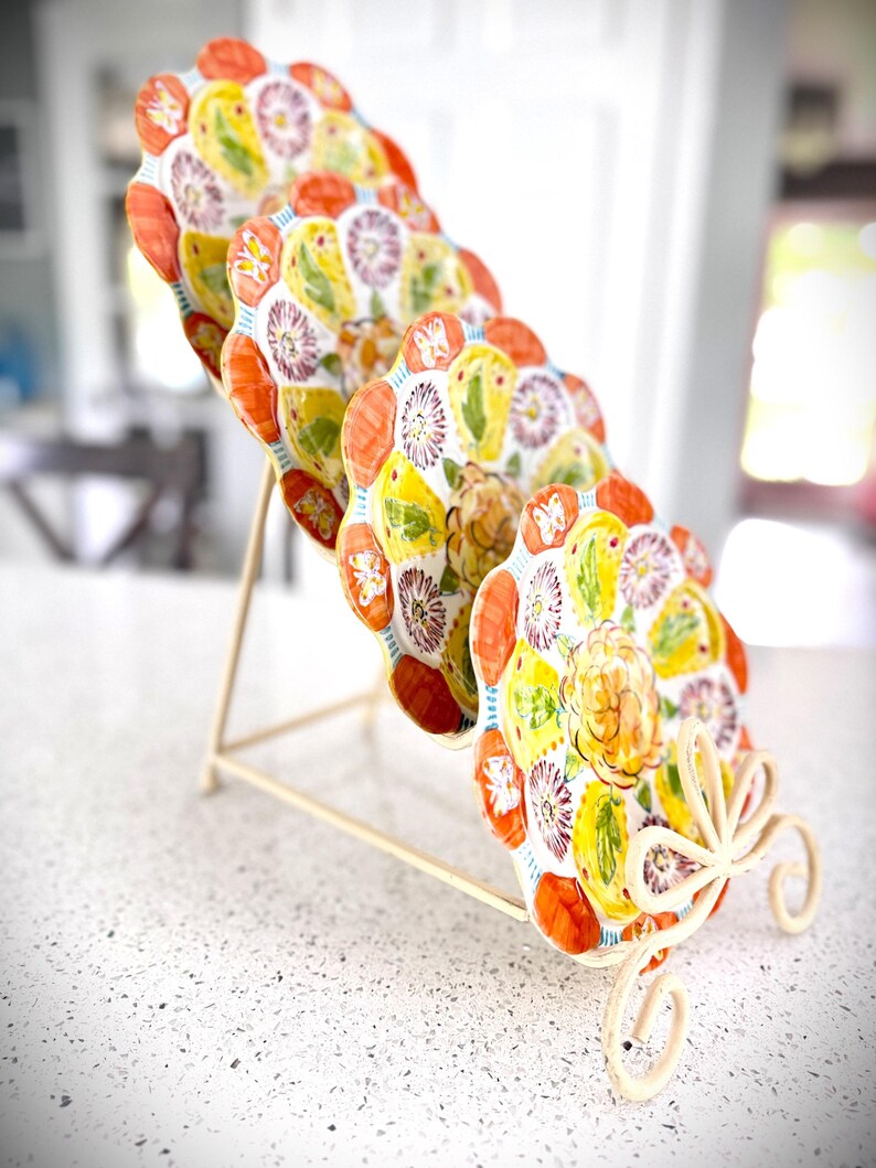 May include: A set of six decorative ceramic plates with a floral design, displayed on a cream-colored metal stand. The plates feature a white background with yellow, orange, and purple floral patterns. The stand has a decorative, scrolled base.