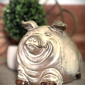 May include: A ceramic piggy bank with a round, stout body and a glossy, cream-colored glaze featuring brown undertones. The pig has a smiling expression with defined facial folds, pointed ears, and dark brown painted hooves on its front feet.