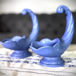 May include: Two matching blue ceramic decorative bowls. Each bowl has a scalloped edge and a curved, twisted handle. The bowls sit on a pedestal base. The background is blurred, with a patterned tablecloth in the foreground.