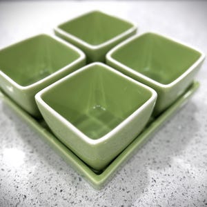 May include: A set of four square, light green ceramic bowls with white rims, arranged on a matching rectangular tray. The bowls are empty and have a glossy finish, perfect for serving snacks or dips.