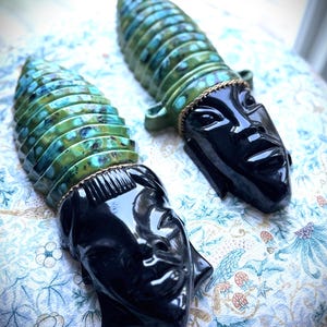 May include: Two ceramic wall plaques featuring stylized human faces with glossy black skin and textured, ribbed headdresses in shades of teal, green, and blue. Each figure is adorned with a thin gold-toned chain detail separating the face and crown.