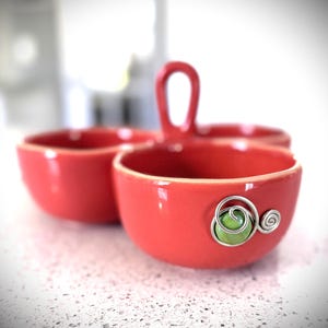May include: A three-sectioned, coral-colored ceramic serving dish with a handle. Each section is a small bowl. One bowl features a green bead and silver wire embellishment. The dish sits on a white speckled surface.