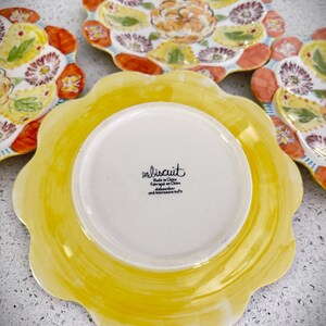 May include: A set of decorative ceramic plates with a floral design. The plates feature a scalloped edge and a vibrant color palette of yellow, orange, and purple. The underside of one plate shows the text "biscuit".