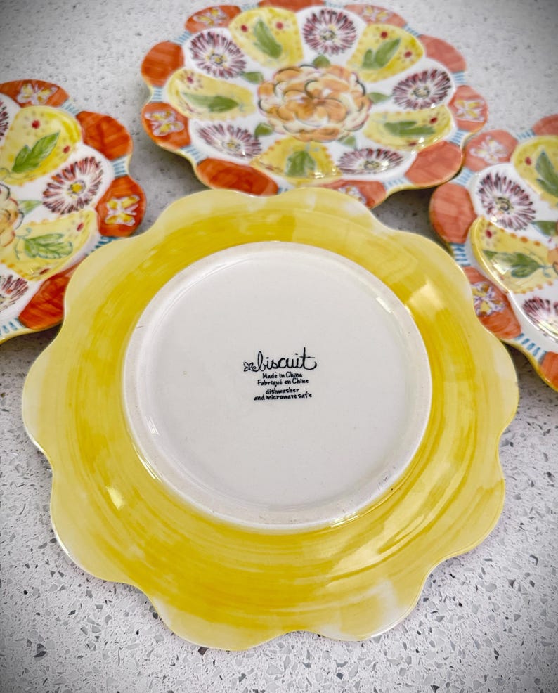 May include: A set of decorative ceramic plates with a floral design. The plates feature a scalloped edge and a vibrant color palette of yellow, orange, and purple. The underside of one plate shows the text "biscuit".
