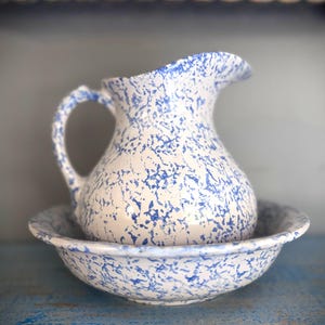 May include: A ceramic pitcher and matching basin set featuring a white base with a blue speckled pattern. The pitcher has a curved handle and a pouring spout, while the basin is round with a wide, flared rim, both displaying a consistent finish.
