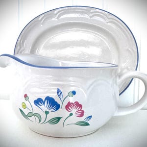 May include: A cream-colored ceramic gravy boat and matching saucer featuring a blue rim and a floral pattern with blue, pink, yellow, and green accents. The gravy boat has a curved handle and a pouring spout, while the saucer has a scalloped edge.