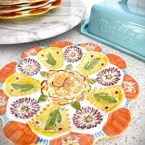 May include: A decorative ceramic plate with a floral design. The plate features a central orange flower surrounded by yellow and orange petal-shaped sections, each with a floral or leaf motif. A stack of similar plates and a blue butter dish are in the background.