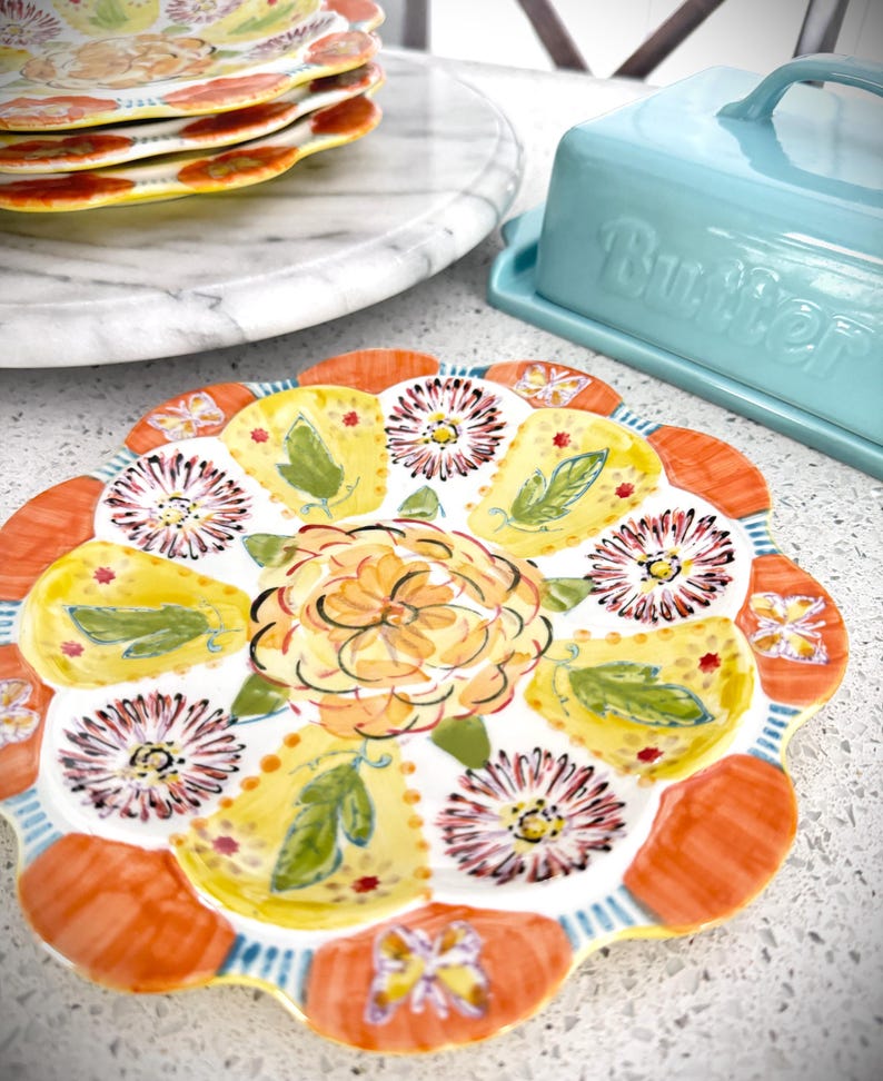 May include: A decorative ceramic plate with a floral design. The plate features a central orange flower surrounded by yellow and orange petal-shaped sections, each with a floral or leaf motif. A stack of similar plates and a blue butter dish are in the background.
