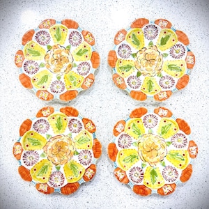 May include: Four decorative plates with a floral design. Each plate features a yellow center with an orange flower, surrounded by a ring of purple flowers, green leaves, and orange scalloped edges. The plates are arranged on a white surface.