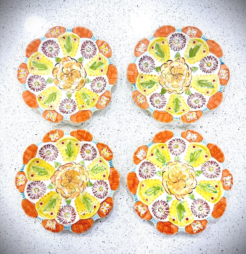 May include: Four decorative plates with a floral design. Each plate features a yellow center with an orange flower, surrounded by a ring of purple flowers, green leaves, and orange scalloped edges. The plates are arranged on a white surface.