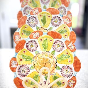 May include: A stack of decorative plates with a floral design. The plates feature a scalloped edge with orange, yellow, and white floral patterns. The plates are held by a white metal stand with a decorative bow.