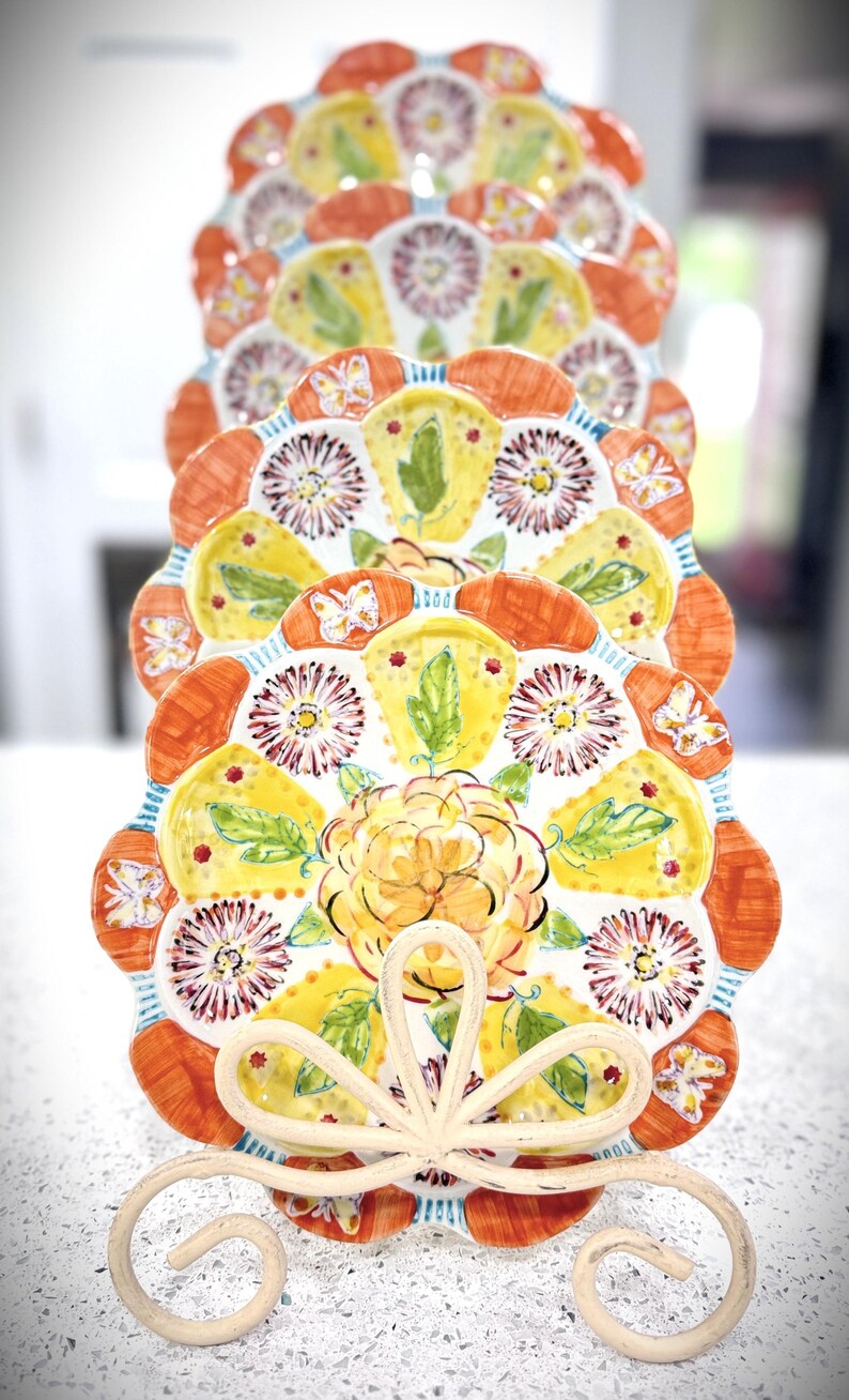 May include: A stack of decorative plates with a floral design. The plates feature a scalloped edge with orange, yellow, and white floral patterns. The plates are held by a white metal stand with a decorative bow.