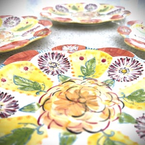 May include: A collection of decorative ceramic plates with a floral design. The plates feature a white base with yellow, red, and purple floral patterns. The edges of the plates have a scalloped design.