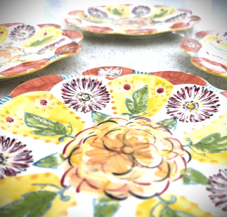 May include: A collection of decorative ceramic plates with a floral design. The plates feature a white base with yellow, red, and purple floral patterns. The edges of the plates have a scalloped design.