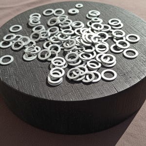 Puede incluir: Una colección de pequeñas arandelas plateadas esparcidas sobre un bloque de madera redondo y oscuro. Las arandelas son circulares con un agujero en el centro y varían ligeramente de tamaño. El bloque tiene una superficie rugosa y texturizada.