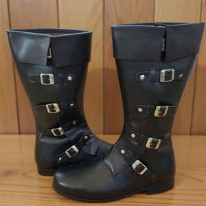 以下が含まれることがあります： 複数のシルバーバックルとストラップが付いた、黒い革製のニーハイブーツのペア。 ブーツは折り返しのトップと丸いつま先が付いています。 ブーツは木製の表面に表示されています。