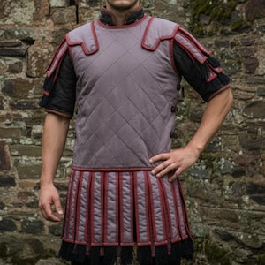 May include: A quilted, gray Roman armor vest with red trim and black sleeves. The vest features a diamond pattern and a pleated skirt with red stripes and black fringe. The armor is designed for historical reenactment or costume use.