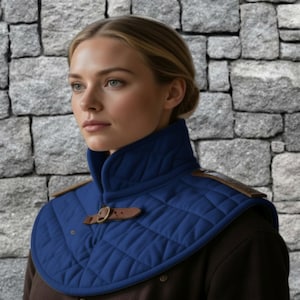 May include: A quilted blue neck guard with brown leather accents and a buckle closure. The neck guard has a high collar and is worn over a brown garment. The background is a stone wall.