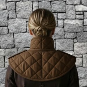 May include: A brown quilted shoulder cape with a high collar. The cape has a diamond pattern and is worn over a dark-colored garment. The background is a stone wall.