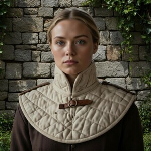 May include: A beige quilted neck piece with a brown leather buckle and shoulder accents. The neck piece is worn over a brown garment. The background features a stone wall and greenery.