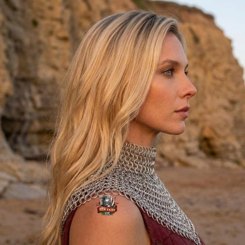 May include: A woman with long blonde hair wearing a silver chainmail collar and a burgundy garment. A small tattoo with the text "Ren Faire US" is visible on the shoulder. The background shows a blurred, natural landscape.
