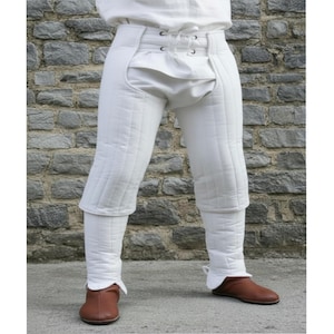 May include: White padded medieval breeches with a high waist and lace-up closure. The breeches have a quilted design and are worn with brown leather shoes. The garment appears to be part of a historical costume or armor.