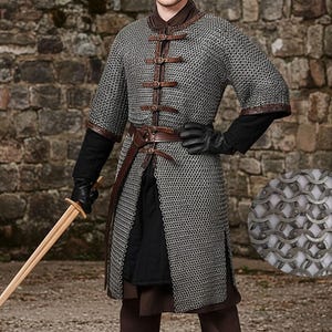 May include: A medieval-style chainmail shirt with leather accents and a brown leather belt. The shirt has short sleeves and is fastened with leather straps. The person is holding a wooden sword. The background is a stone wall.