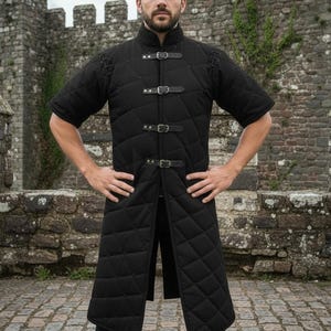 May include: Black quilted medieval gambeson with short sleeves and leather buckle closures. The long coat features a diamond pattern and a high collar. The garment is designed for historical reenactment or costume use.