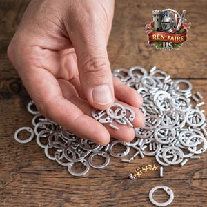 May include: A hand holding a few small, silver-colored metal keychains, with many more scattered on a wooden surface. The keychains have a circular design with a key-shaped element. The image includes a logo that says "REN FAIRE US".