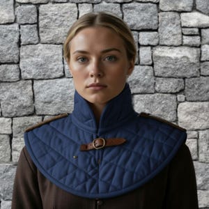 May include: A quilted navy blue neck guard with brown leather accents and a buckle closure. The neck guard has a diamond pattern and is worn over a brown garment. The background is a stone wall.