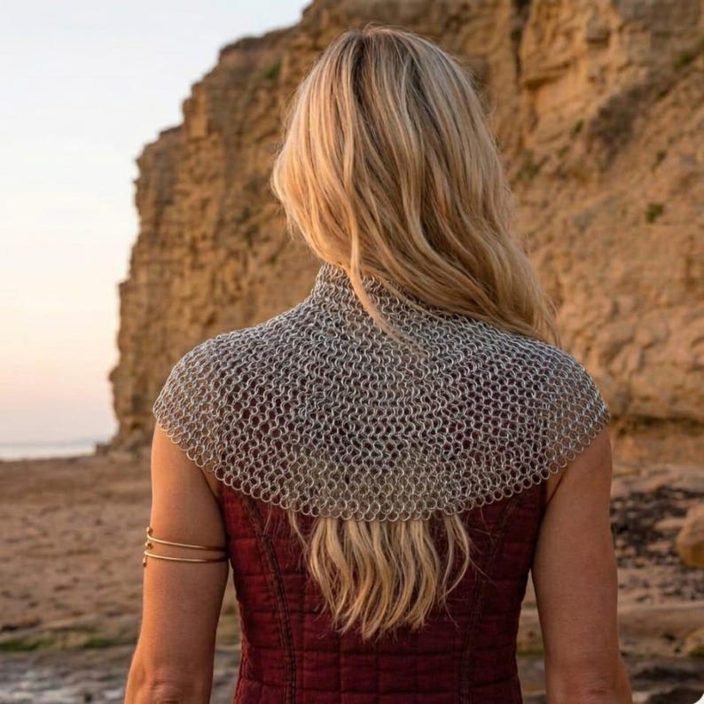 May include: A silver chainmail cowl over a burgundy top. The chainmail is made of small, interlocking metal rings. The person is facing away from the camera, with long blonde hair. A gold arm cuff is visible.