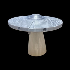 May include: A light gray, 3D-printed model of a UFO, featuring a disc-shaped top with radial lines and three circular openings. The UFO rests on a translucent, conical base, set against a black background.