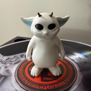 May include: A white creature figurine with large black eyes, small brown horns, and blue-tinted ears standing on a vinyl record. The record label features the text "Fueled By Ramen" and "Breach" in a circular pattern around the center hole.