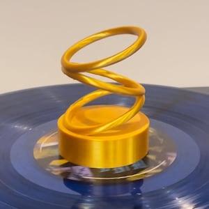 May include: A gold-colored, spiral-shaped decorative object. The spiral sits atop a cylindrical base, resting on a blue vinyl record. The design is modern and abstract, with a glossy finish.
