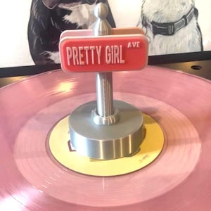 May include: A pink vinyl record playing with a silver record weight. The record weight has a red sign that reads "PRETTY GIRL AVE". The background features two dog portraits.