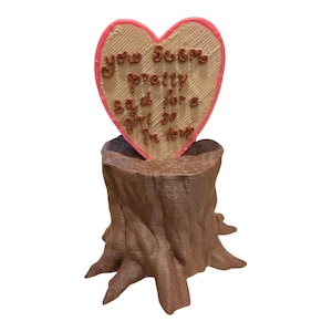 May include: A brown tree stump sculpture supporting a heart-shaped plaque with a pink border. The plaque features the embossed brown text: you seem pretty sad for a girl so in love. The object has a textured finish throughout the entire structure.