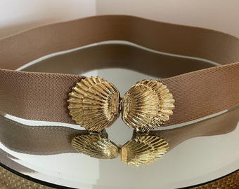 May include: A tan elastic belt with a gold-colored seashell clasp. The belt is displayed on a reflective surface, showcasing the clasp's intricate details and the belt's texture. The clasp is made of two seashell-shaped pieces that connect in the center.