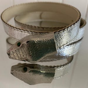 May include: A silver snake-shaped belt with a textured, scaly design. The snake's head has green eyes. The belt coils around itself. The belt is made of a shiny, metallic material. The belt's design is reminiscent of a snake.