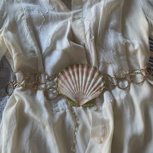 Puede incluir: Una camisa color crema con botones y un cinturón de cadena dorada con una gran concha marina como pieza central. La concha tiene rayas rosas y blancas. El cinturón está drapeado sobre la camisa.