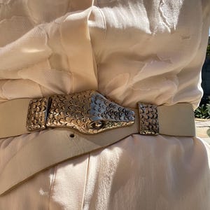 Puede incluir: Cinturón de cuero color crema con hebilla de cabeza de serpiente dorada. El cinturón se lleva sobre una blusa color crema. La hebilla de la cabeza de serpiente está detallada con escamas y un acabado texturizado. El cinturón mide unos 3,8 cm de ancho.