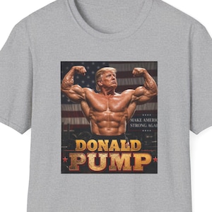 Peut inclure: T-shirt gris clair avec un imprimé d'un homme musclé avec l'inscription dorée "DONALD PUMP". Le motif comprend un drapeau américain et les mots "MAKE AMERICA STRONG AGAIN".