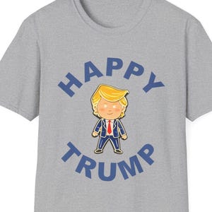 K&ouml;nnte beinhalten: Ein graues T-Shirt mit einer Cartoon-Illustration einer Person mit blonden Haaren und einem blauen Anzug. Die Worte "HAPPY TRUMP" sind in Blau gedruckt und w&ouml;lben sich &uuml;ber und unter der Illustration.