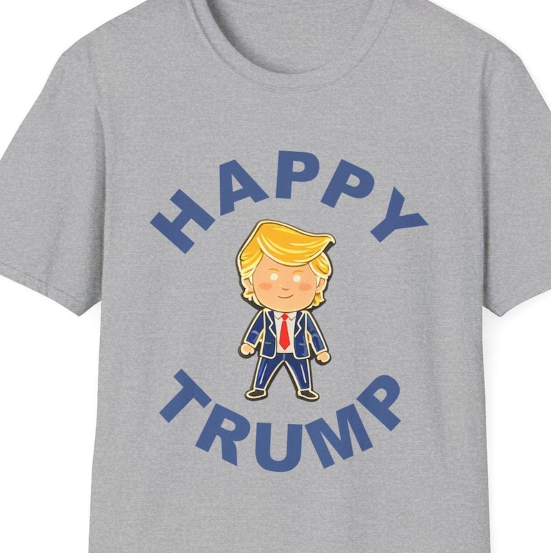K&ouml;nnte beinhalten: Ein graues T-Shirt mit einer Cartoon-Illustration einer Person mit blonden Haaren und einem blauen Anzug. Die Worte "HAPPY TRUMP" sind in Blau gedruckt und w&ouml;lben sich &uuml;ber und unter der Illustration.