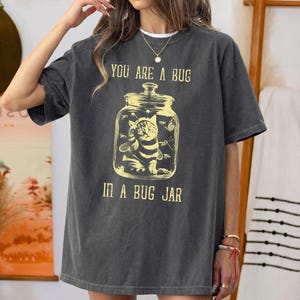 Op de afbeelding: Een antracietgrijs t-shirt met korte mouwen en een gele grafische print van een kat met bijenvleugels in een glazen pot. De tekst boven de pot luidt YOU ARE A BUG en de tekst onder de pot luidt IN A BUG JAR in een verweerd lettertype.