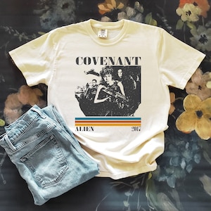 Puede incluir: Camiseta color crema con un diseño gráfico de la película "Alien: Covenant". El diseño incluye la palabra "COVENANT" sobre una imagen en blanco y negro de personajes, con "ALIEN" y "2017" debajo. Vaqueros de mezclilla azul claro doblados junto a la camiseta.