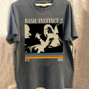 May include: Dark gray t-shirt featuring a graphic from the movie "Basic Instinct 2." The design includes the movie title, a black and white image of a woman, and the year 2006. The shirt has a crew neck and short sleeves.